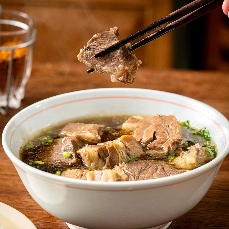 Chopsticks lifting a piece of tender beef from a steaming bowl of Thai beef noodle soup.