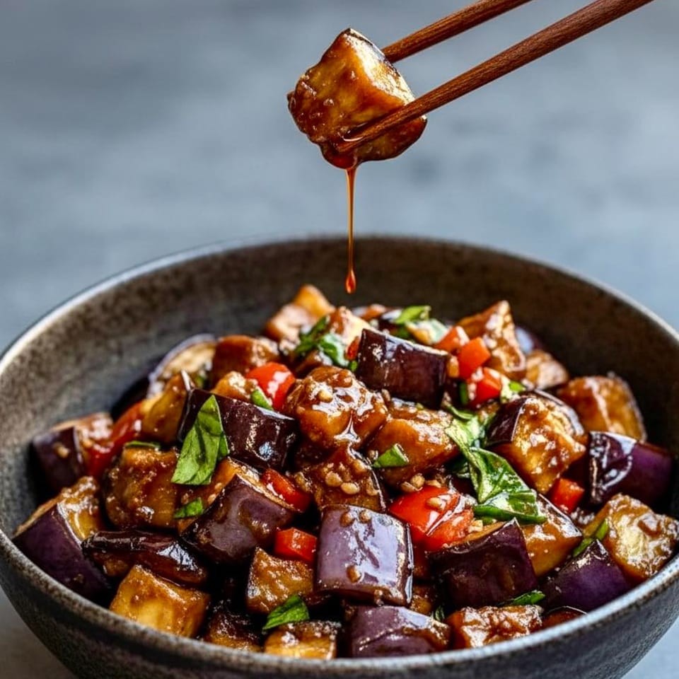 Chopsticks holding a piece of glazed eggplant over a bowl of stir-fried eggplant, peppers, and basil.