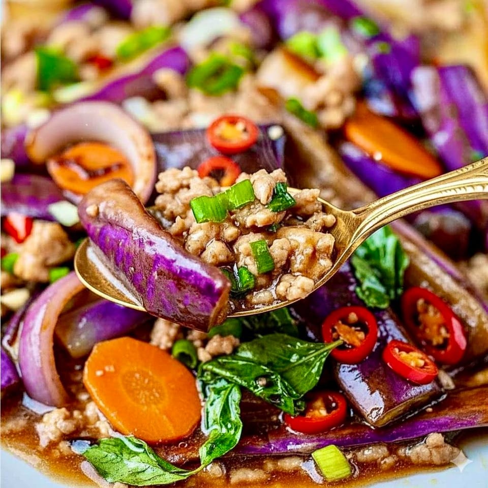 A golden spoon lifts a piece of purple eggplant and ground meat from a Thai dish.